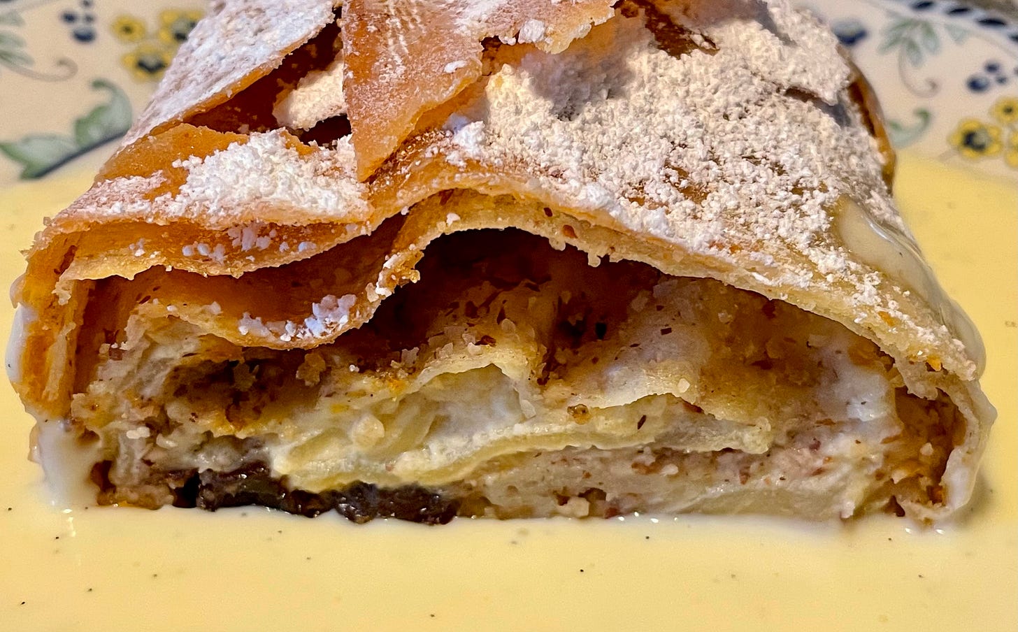 Close-up of a slice of Apfelstrudel with golden, flaky layers, filled with apples, nuts, and raisins, topped with powdered sugar, and served on a plate with vanilla sauce. Close-up of a slice of Apfelstrudel with golden, flaky layers, filled with apples, nuts, and raisins, topped with powdered sugar, and served on a plate with vanilla sauce.