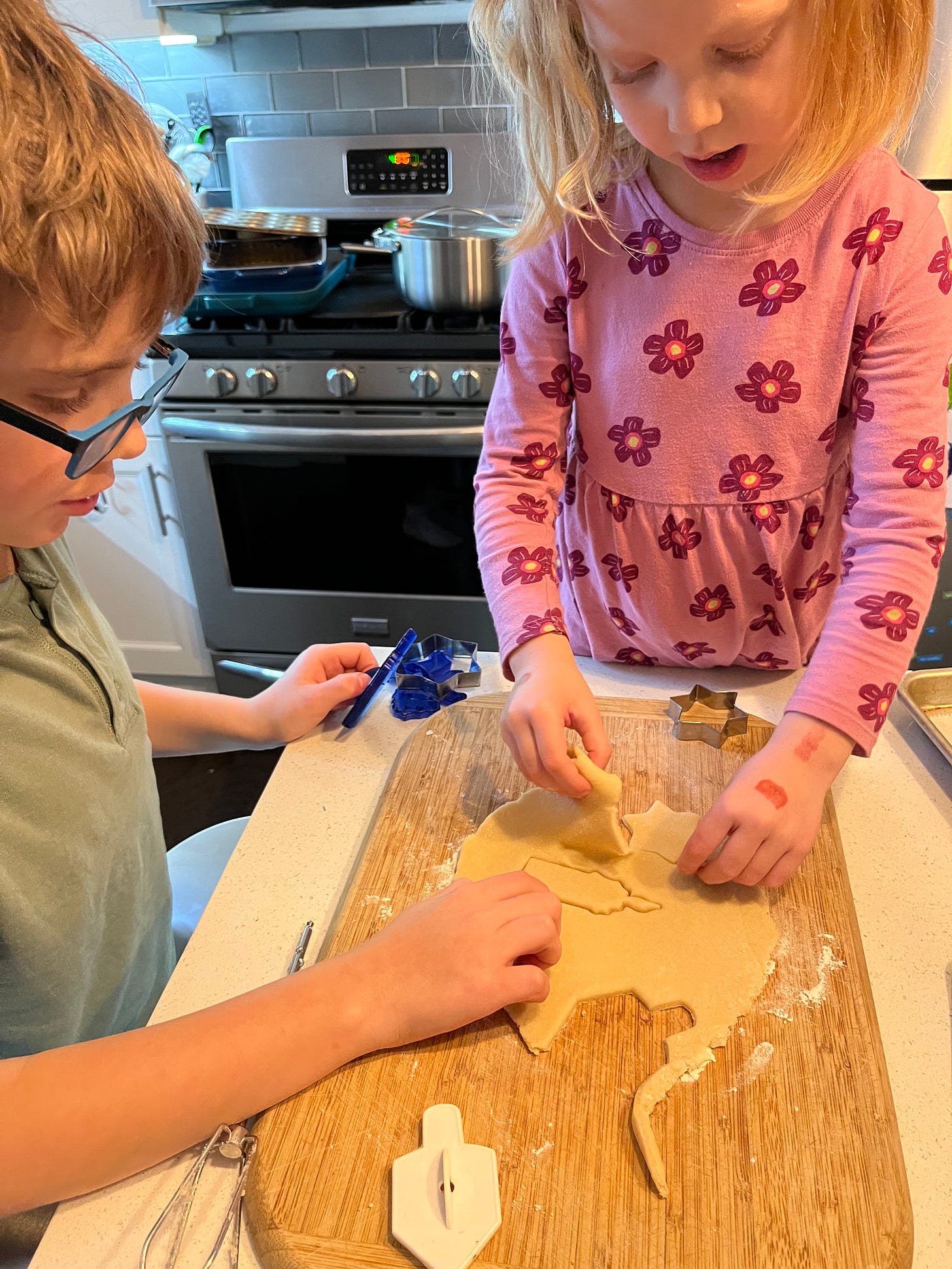 A young boy and young girl cutting out Hanukkah-themed sugar cookies at a kitchen counter on a wooden cutting board.