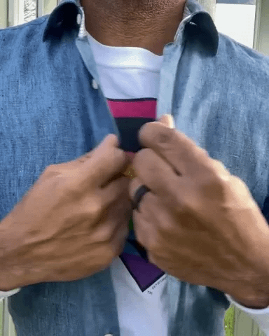 Video gif. We zoom in on a man's chest as he opens a collared shirt to reveal a rainbow Superman shield on a t-shirt underneath.