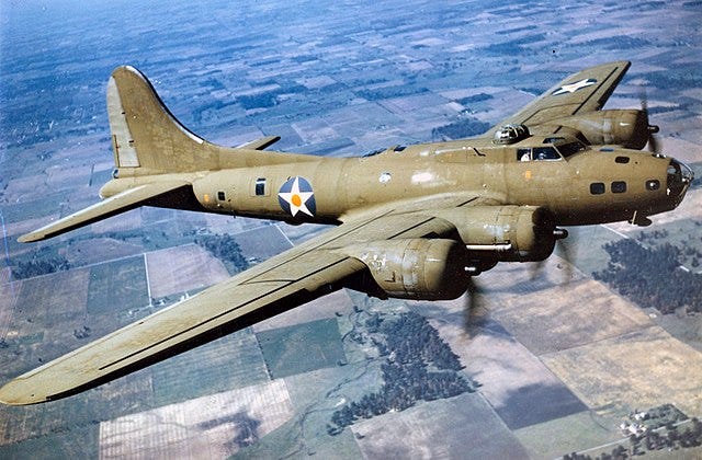 A World War II-era Boeing B-17E Flying Fortress bomber in flight over a patchwork of green and brown fields, showcasing its olive drab paint, U.S. Army Air Forces insignia, and distinctive four-engine configuration.