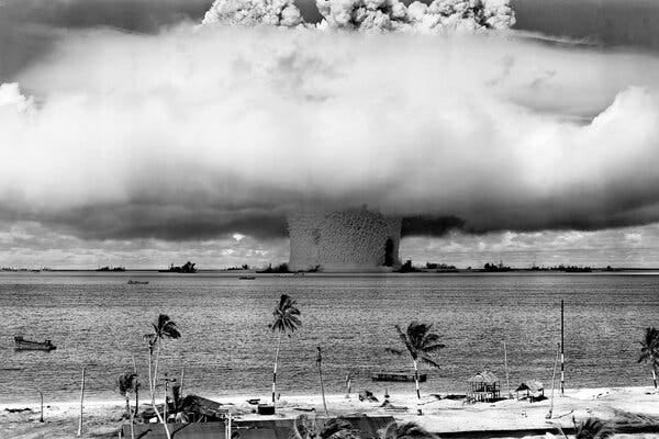 In a black and white picture, a mushroom cloud rises out of the sea during a nuclear bomb test.