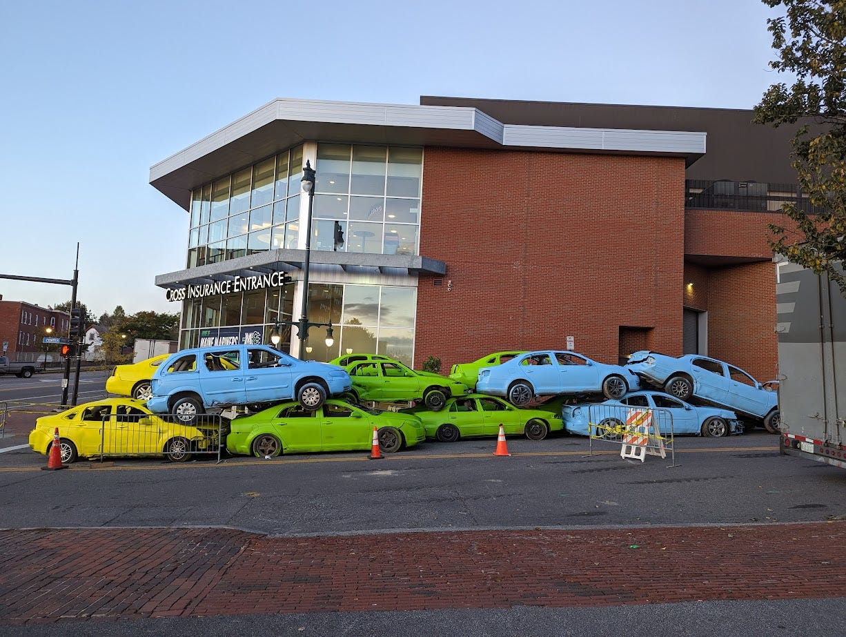 A bunch of old cars, some piled two high, which are all monochromatically painted. Some are bright yellow, some are sky blue, some are bright spring green. They are on a street in Portland, Maine.