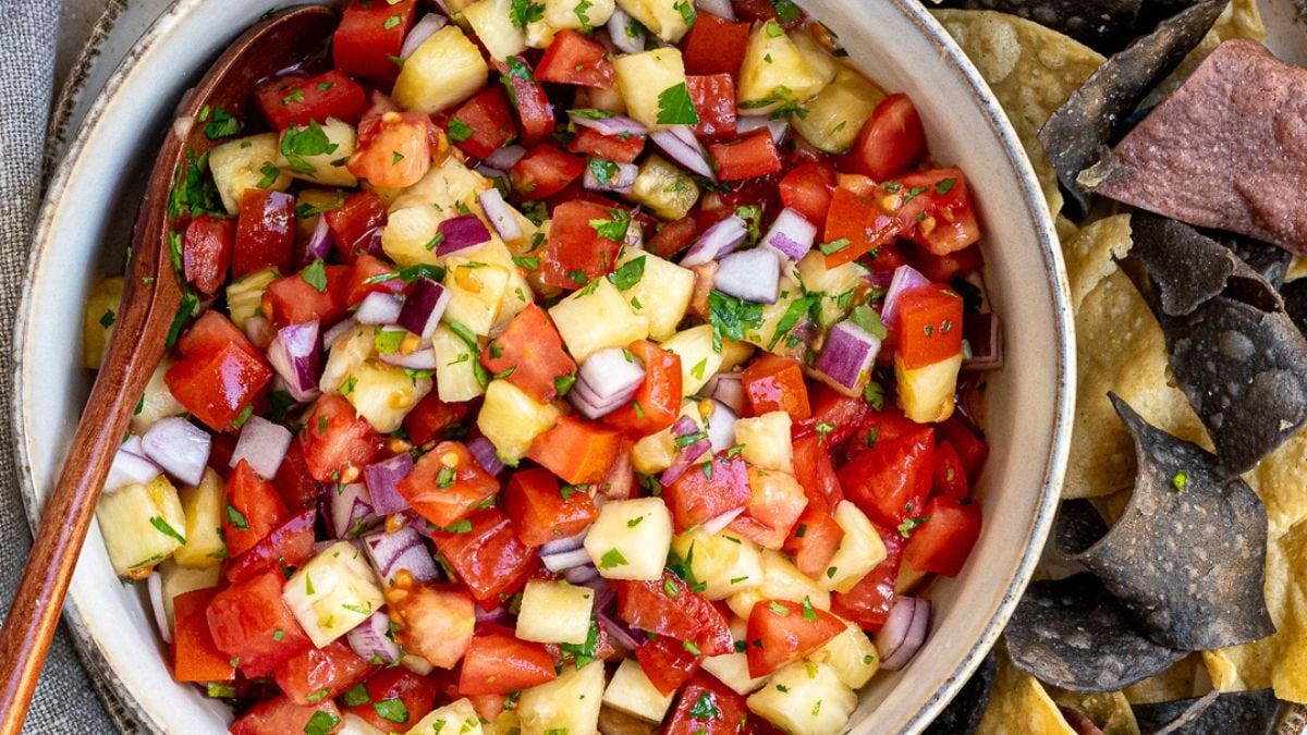A colorful bowl of diced tomato, pineapple, and red onion salsa garnished with chopped cilantro, served with a side of multicolored tortilla chips.