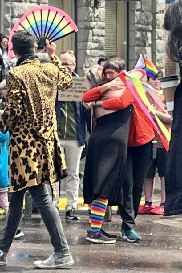 To the left, a young person in a leopard print jacket, jeans,and high top tennis shoes carries a rainbow fan down a rainy street. He is looking at the people on the right, a young woman in a black dress wearing rainbow socks and carrying a rainbow flag, hugging a woman along the side of the street wearing a red raincoat.