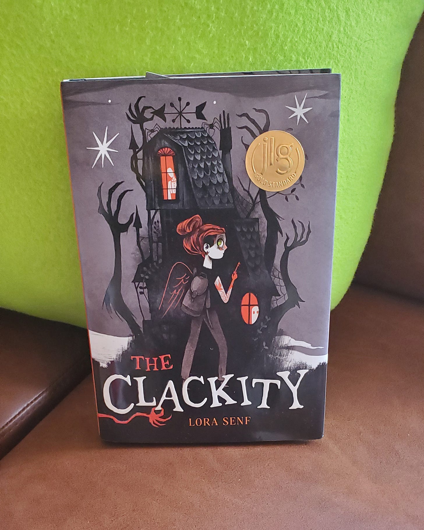 A young girl with red hair and light skin with a backpack and key in her hand walks in front of a black two-story house, with arms coming out of the house referring to the book’s title, “The Clackity” by Lora Senf. 