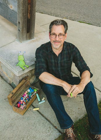 the artist David Zinn seated next to a wooden box of chalk and his first chalk character, Sluggo
