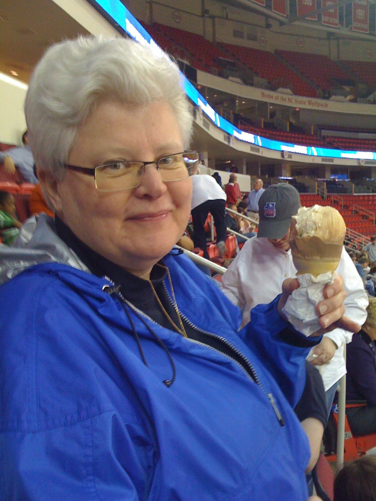 An older woman with white hair, a blue jacket, and holding an ice cream cone in a stadium