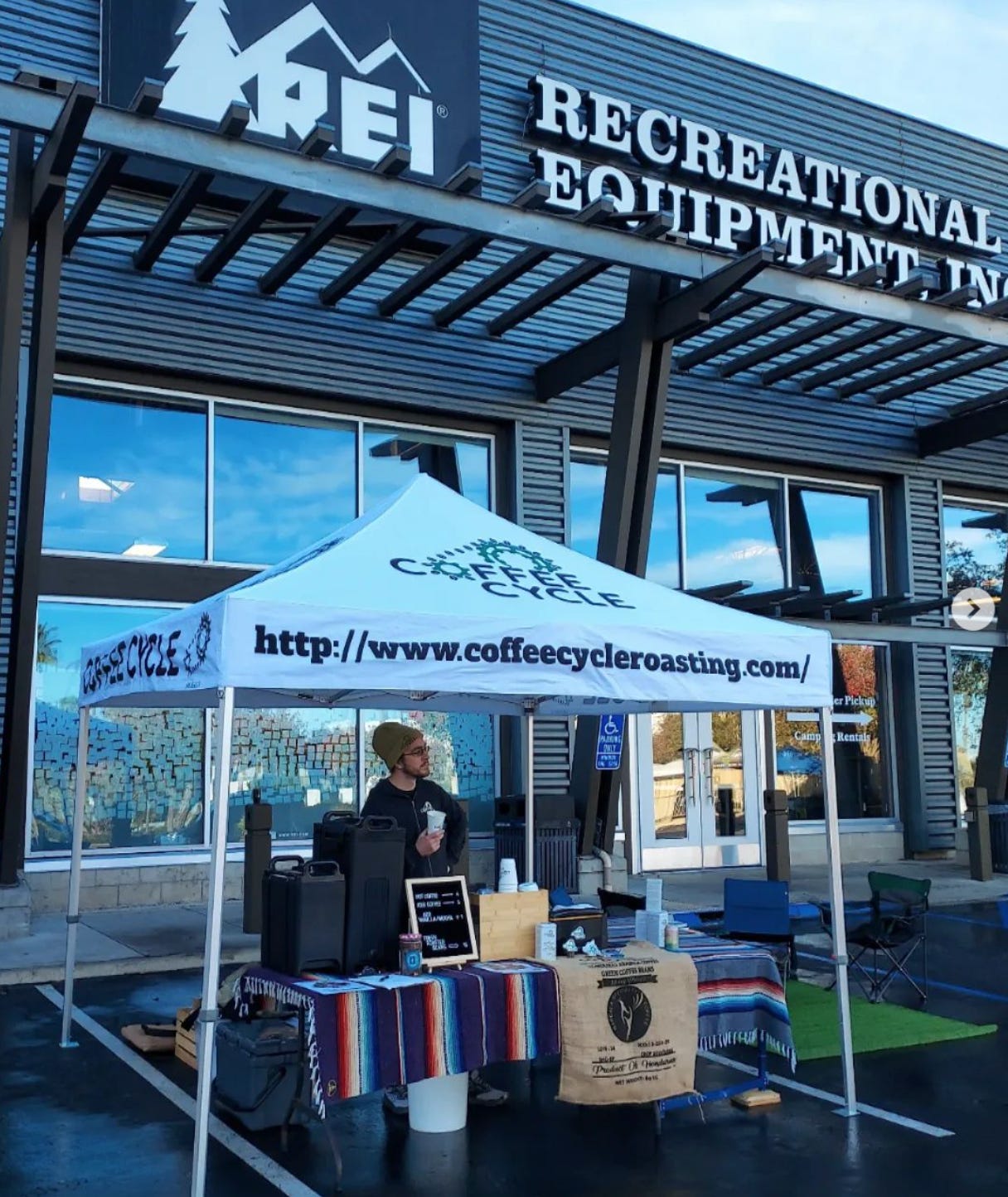 A barista in a hoodie and stocking cap stands behind a folding table covered in coffee dispensers and coffee cups under a pop-up tent in front of an REI store in the early morning.
