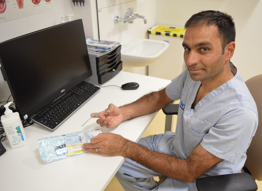 Professor Mahmood Bhutta wearing scrubs, sitting at a desk holding reusable crocodile forceps.