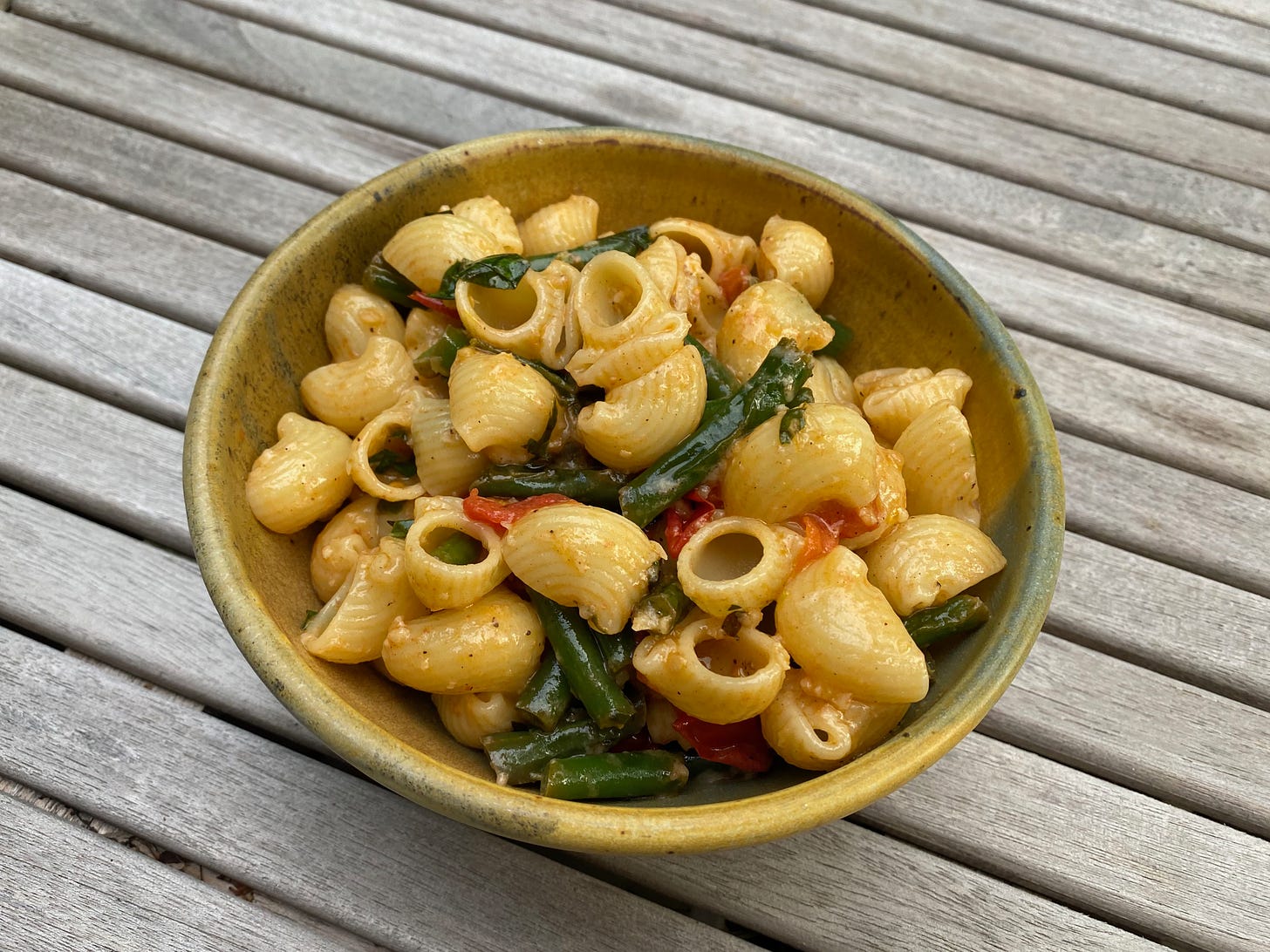 A ceramic bowl of creamy pasta, roasted green beans, and flecks of red tomatoes on a porch table.