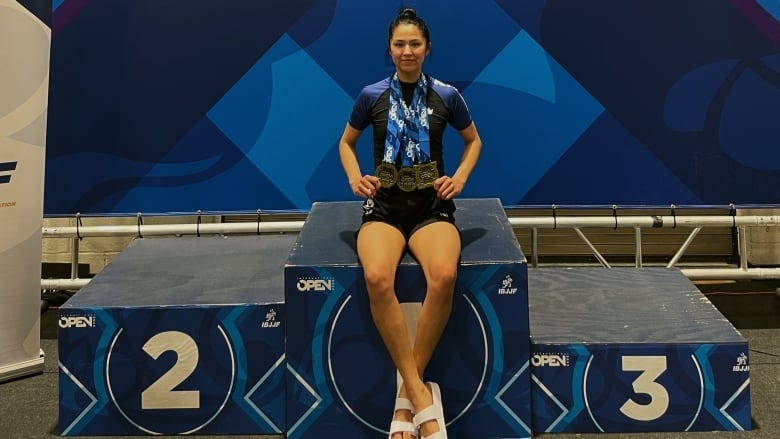 A woman with three bronze medals around her neck sits on a blue multi-layered podium