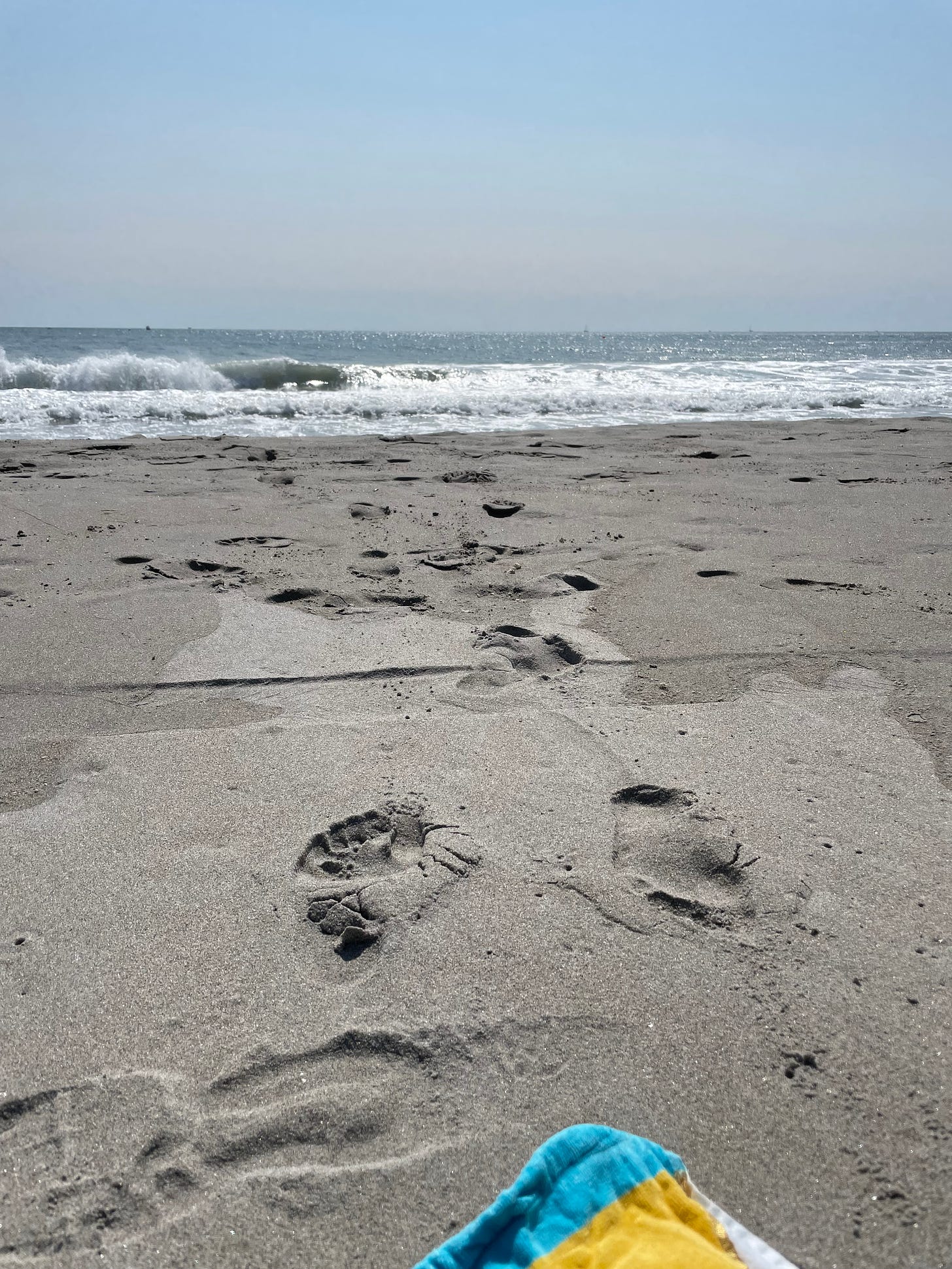 It is a day with blue clear skies. In the background, rushing waves can be seen. In the foreground, the edges of a beach towel can be seen as footprints on a sandy beach lead out to shore.