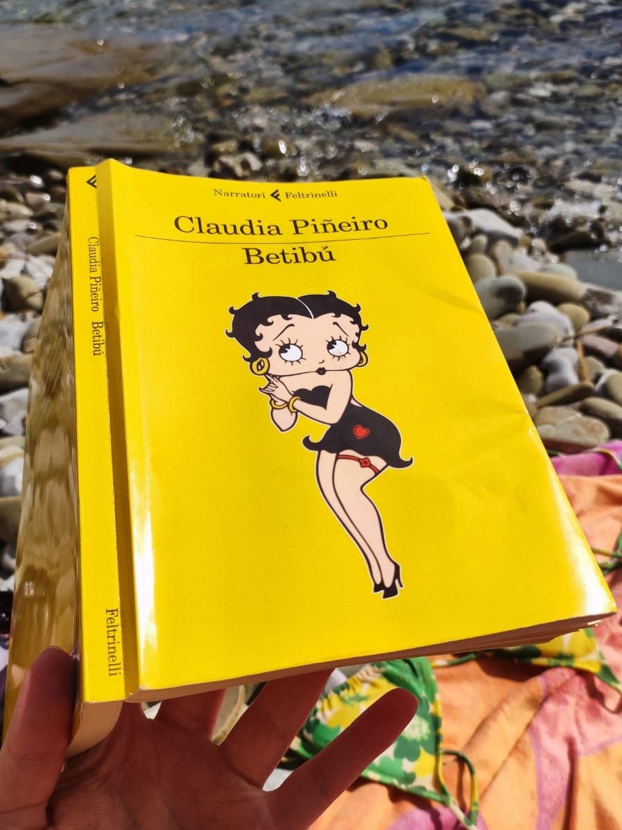 Un libro giallo tenuto in mano. Sullo sfondo sassi, mare e un telo mare rosa.