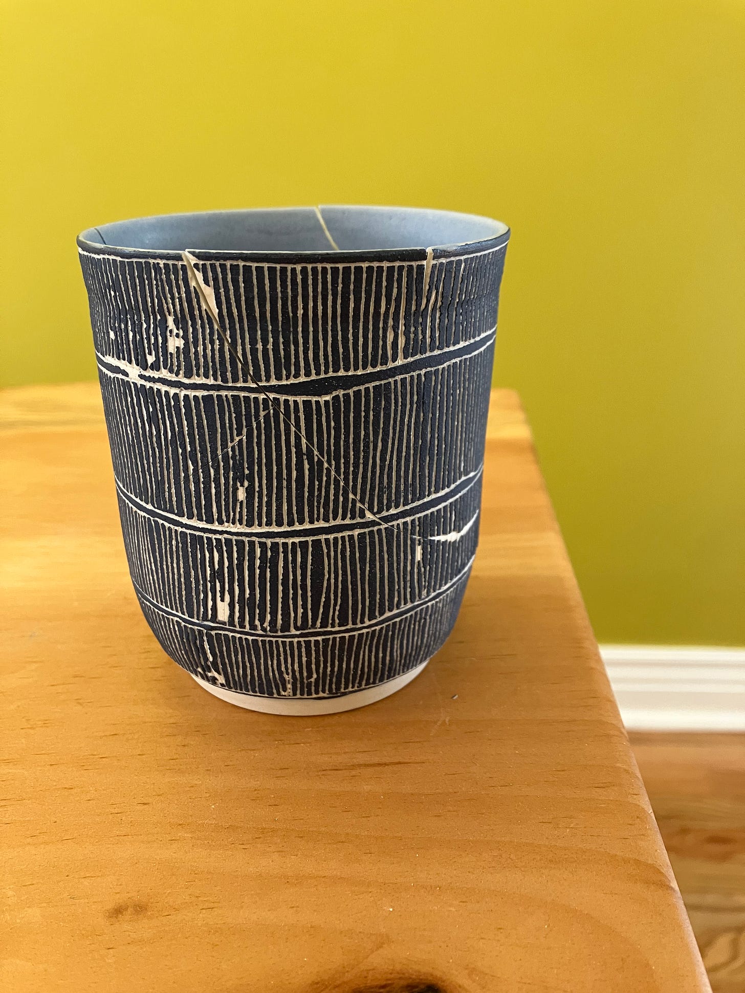 an anxiety cup engraved with dark blue sgraffito and a light blue matte glaze inside. it's been broken and fit back together loosely, on a table, in front of a green background.