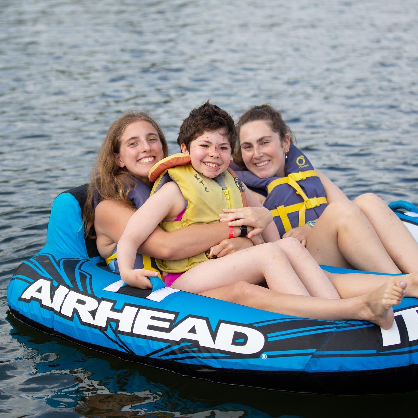 a child sits in an inflatable boat with two camp staff on a lake