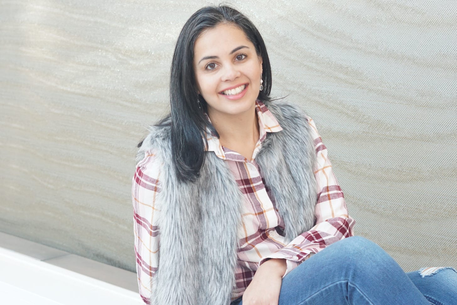 A headshot of Taslim Jaffer, a woman with dark shoulder-length hair smiling at the camera, wearing a plaid shirt, fuzzy grey vest, and jeans
