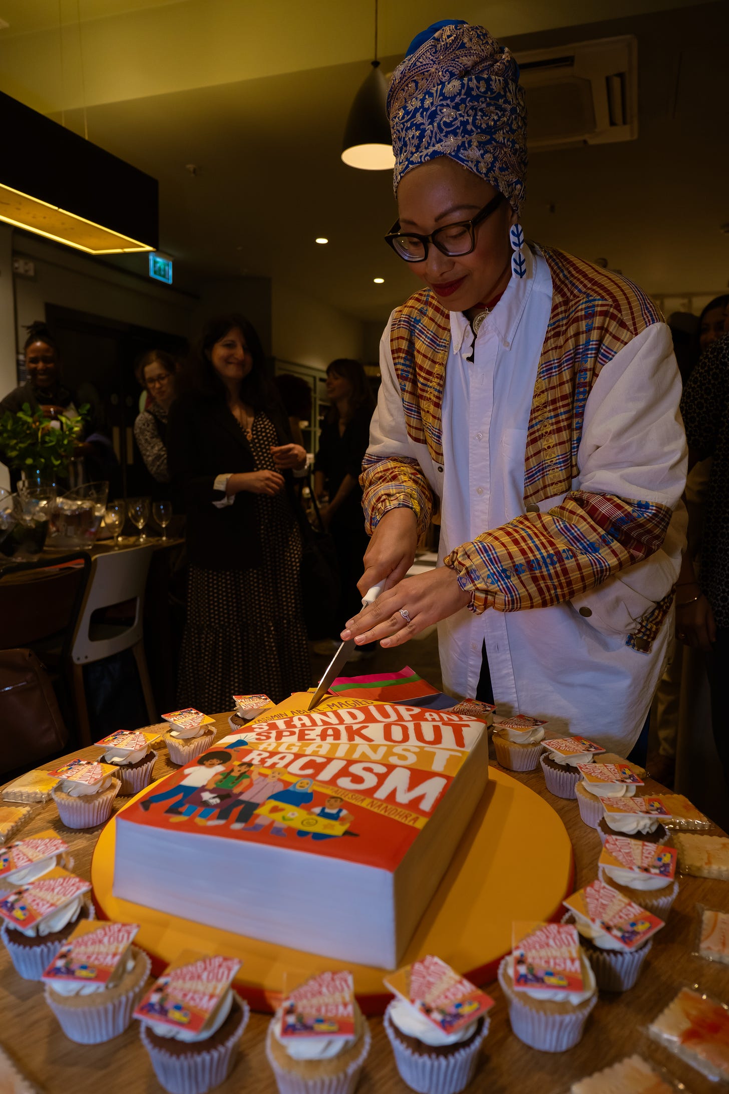 Yassmin Abdel-Magied wearing a blue glittery turban and white jacket with red and gold detailing cutting into a large cake with the cover of Stand Up and Speak Out Against Racism (her book) printed on the cake surface. The cake is surrounded by cupcakes and Yassmin looks pleased.