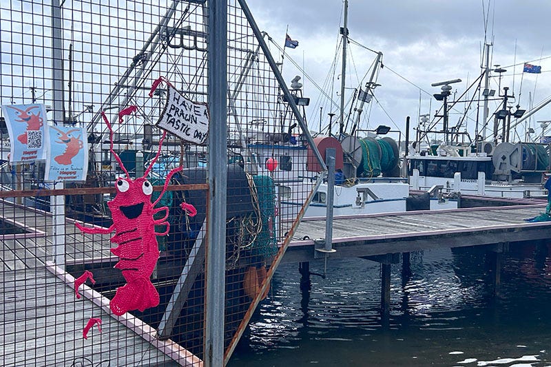 A gate in front of a harbour full of boats. The gate features a crotched prawn and a sign saying 'Have a prawn-tastic day'.