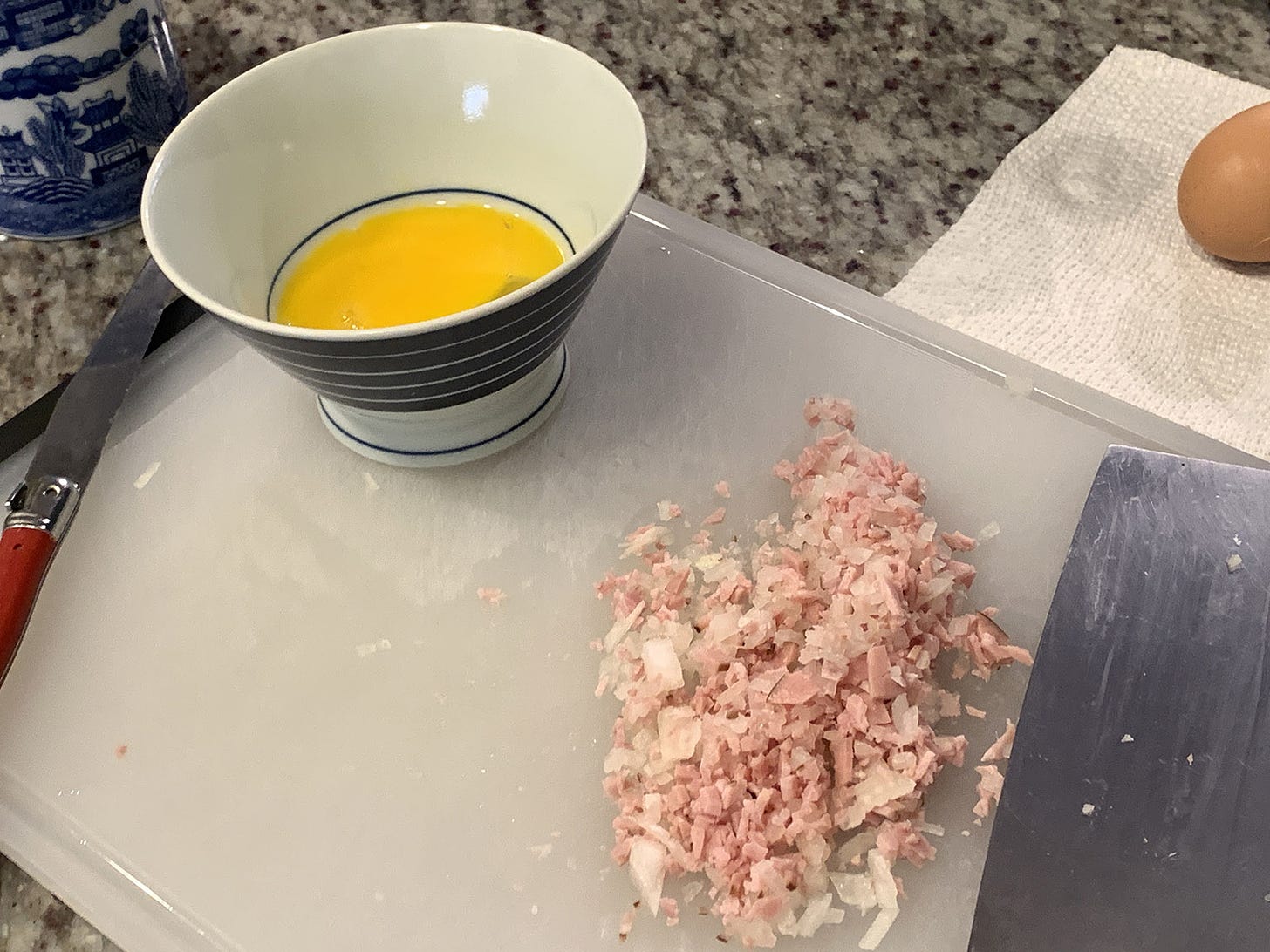 A bowl with a broken egg in it and some chopped ham on a cutting board