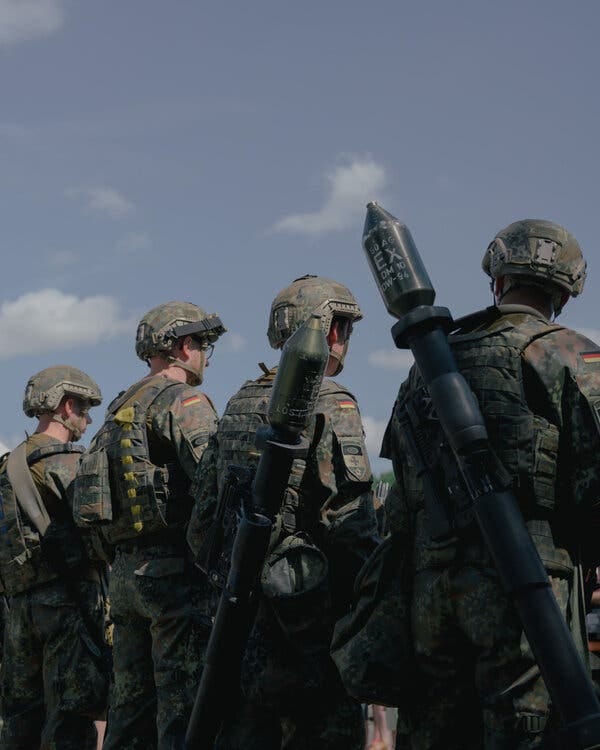 A photograph of four soldiers, two with weapons on their backs, facing away from the camera.