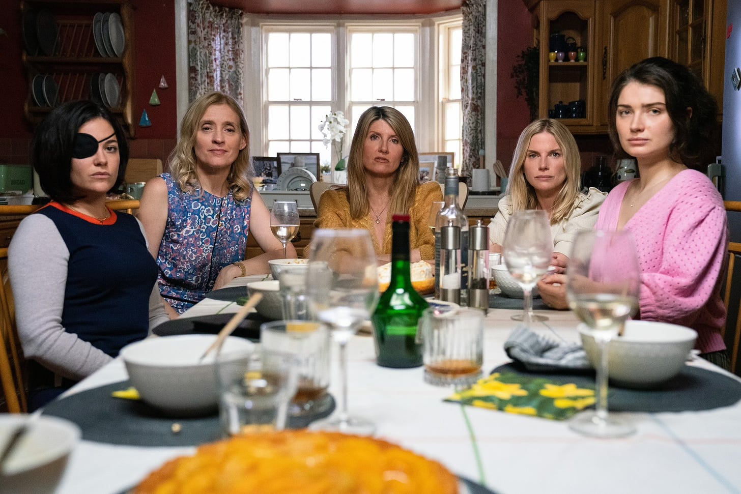 The 5 sisters from Bad Sisters sit at a kitchen table looking expressionless at the camera. Sharon Horgan (Eva) is at the head of the table with 2 sisters flanking her on each side.