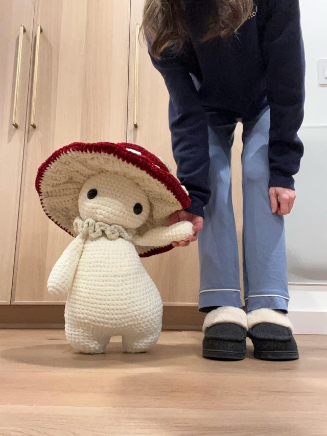 A person, face obscured by their hair, leans down to hold hands with a knee-high crochet mushroom doll.
