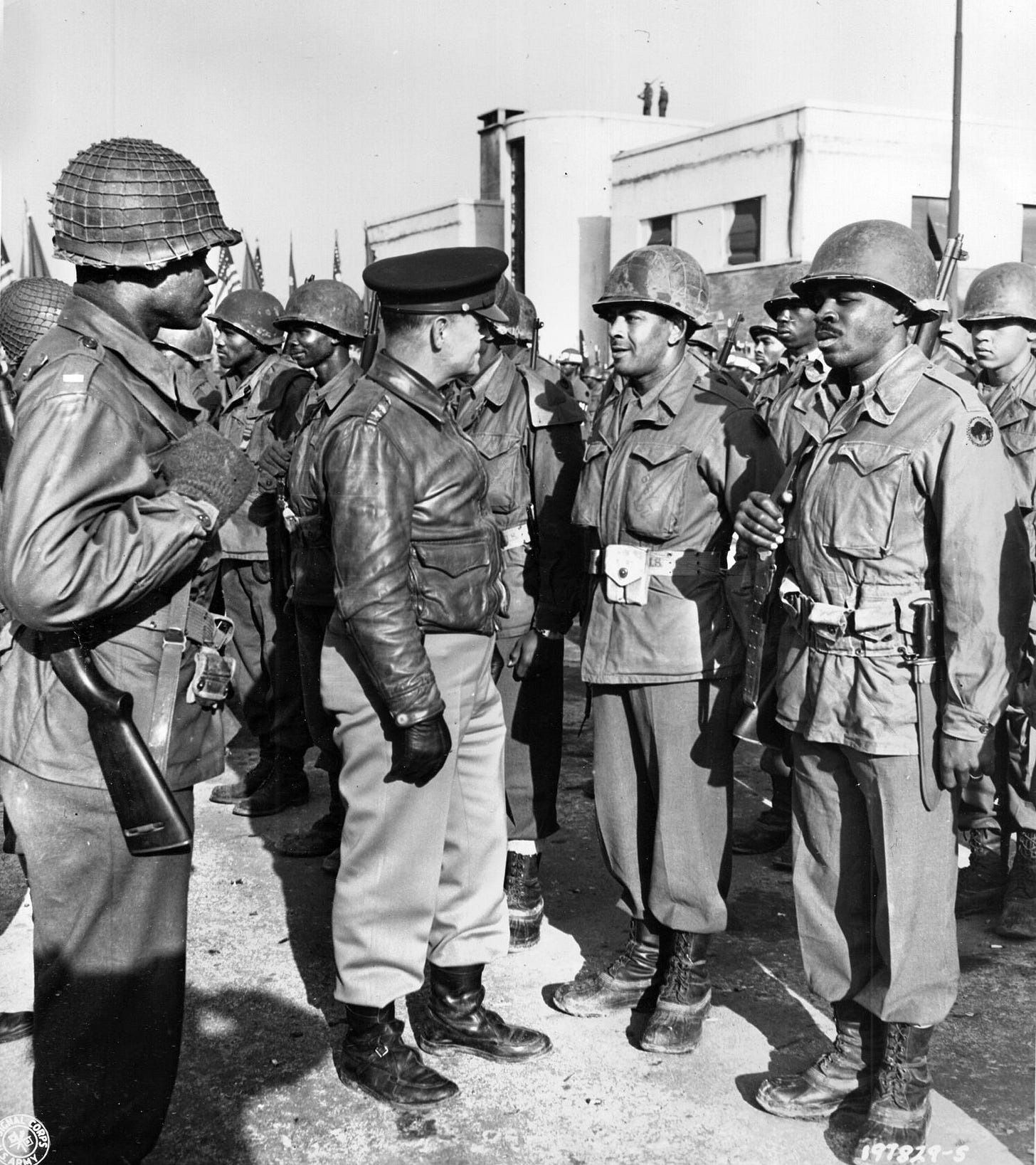 Lieutenant General Lucian K. Truscott, commander of the Allied Fifth Army in Italy, pauses to chat with soldiers of the 92nd Infantry Division after the American troops have stood their ground in defense of the hills above the town of Viareggio. Sentries are alert atop the adjacent buildings.