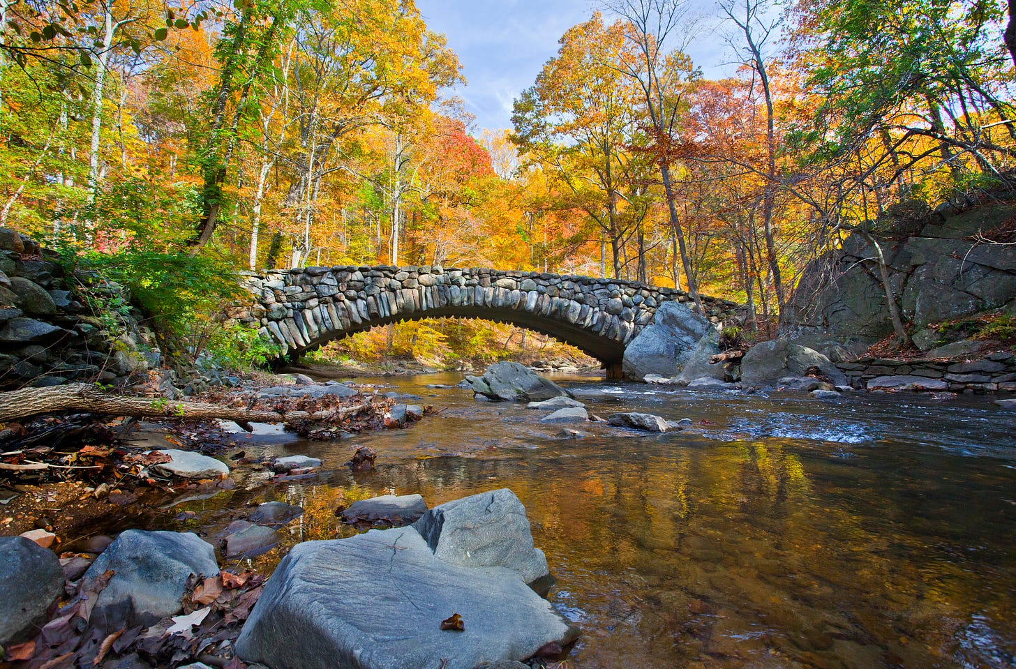A bridge made of rocks stands over a rocky creek in the fall time.