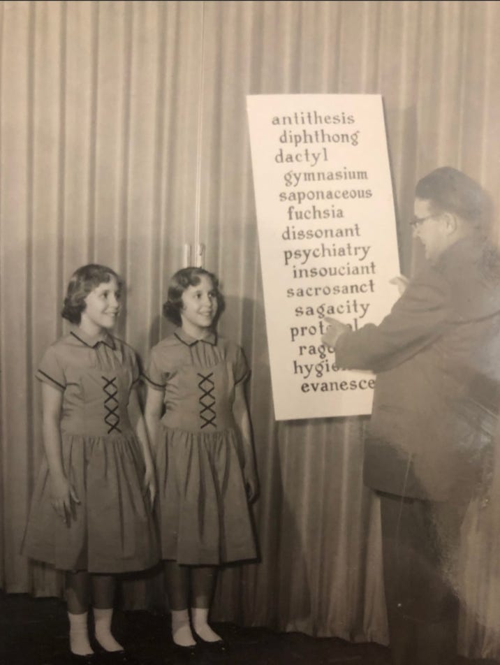 twin girls standing next to a large list of spelling bee words being pointed at by a man