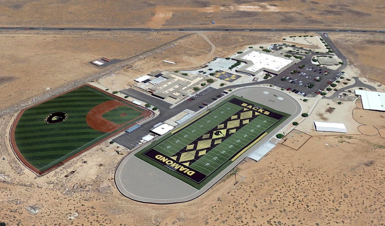 An aerial photograph of a campus, surrounded by desert, featuring a football field with "DIAMOND" written in the grass of one endzone and "BACKS" written in the other, as well as a baseball diamond