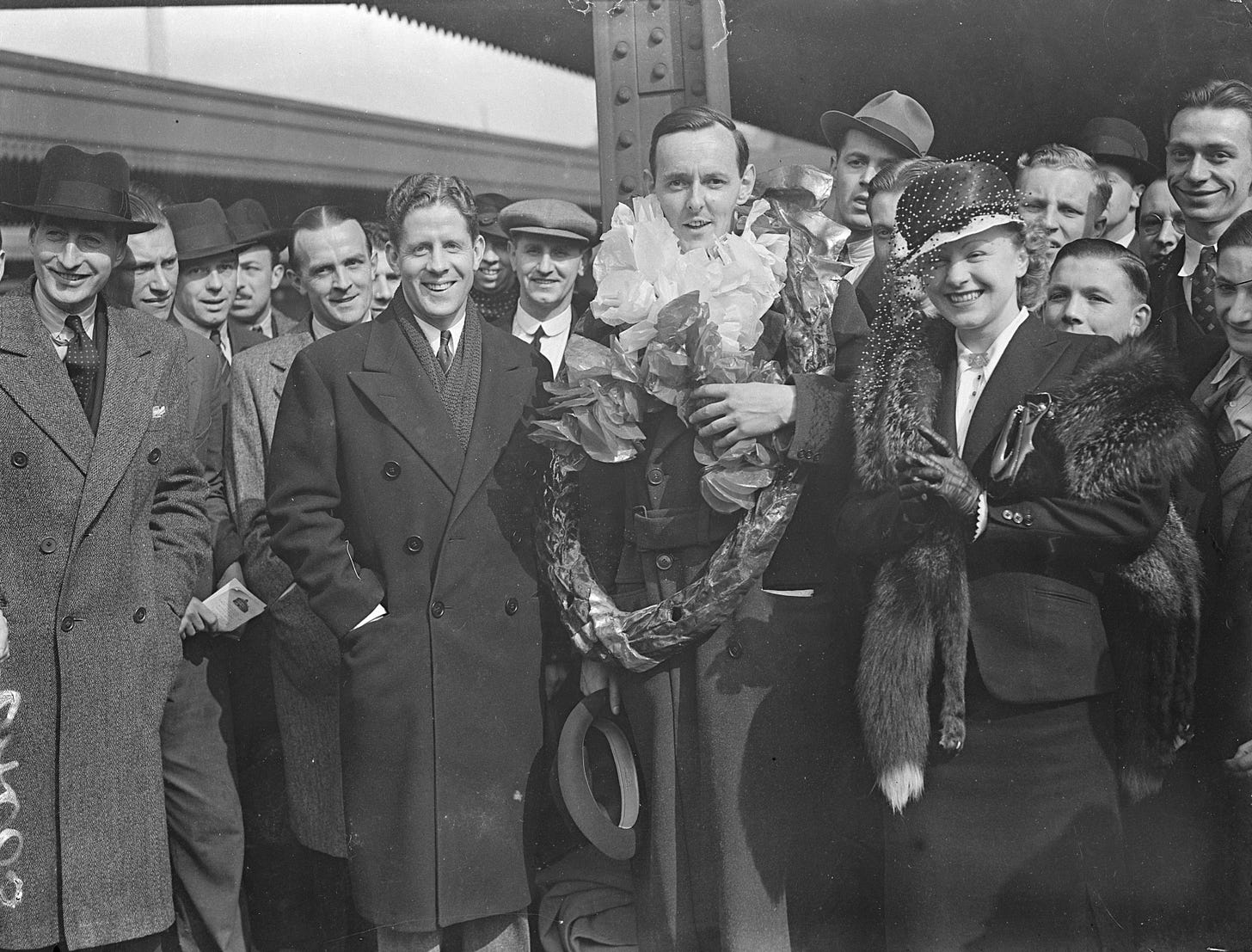 Eric Maschwitz surrounded by a group of people with several paper wreaths around his neck