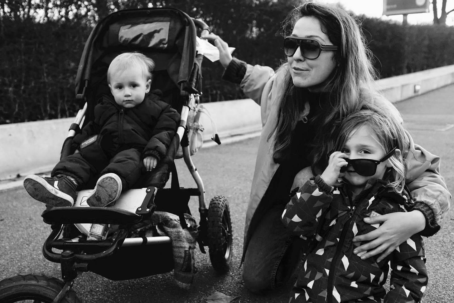 A black and white photo of Ellese looking very cool in sunglasses, with her arm wrapped around Michael, in a big jacket and also wearing sunglasses. Her hand is on the buggy, where Angelo sits, staring eagerly at the camera.