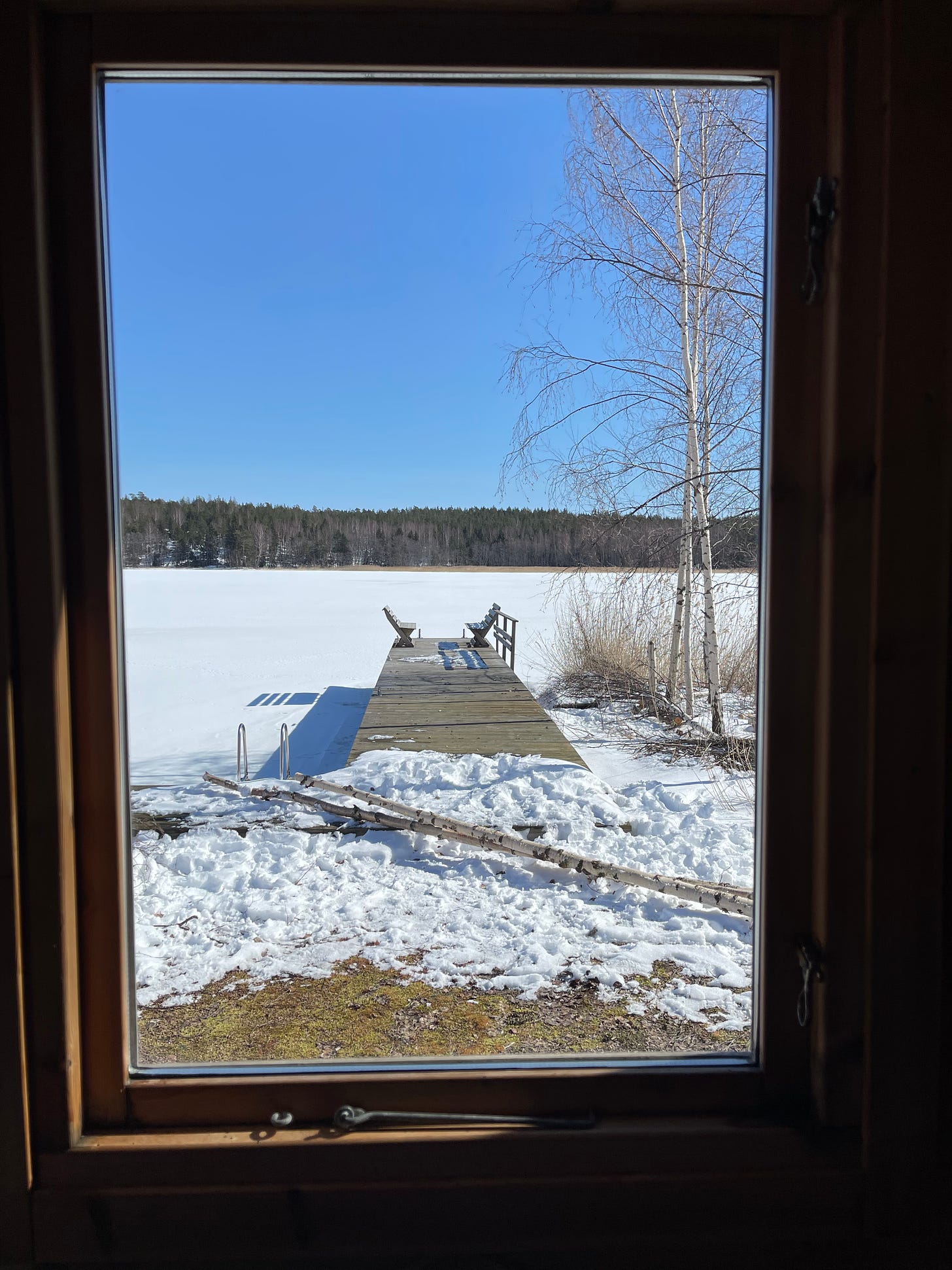 Looking out a rectangular window of the sauna change room to a wood jetty with 2 bench seats at the end, a blue sky and the white, snow covered water and a line of forest in the distance