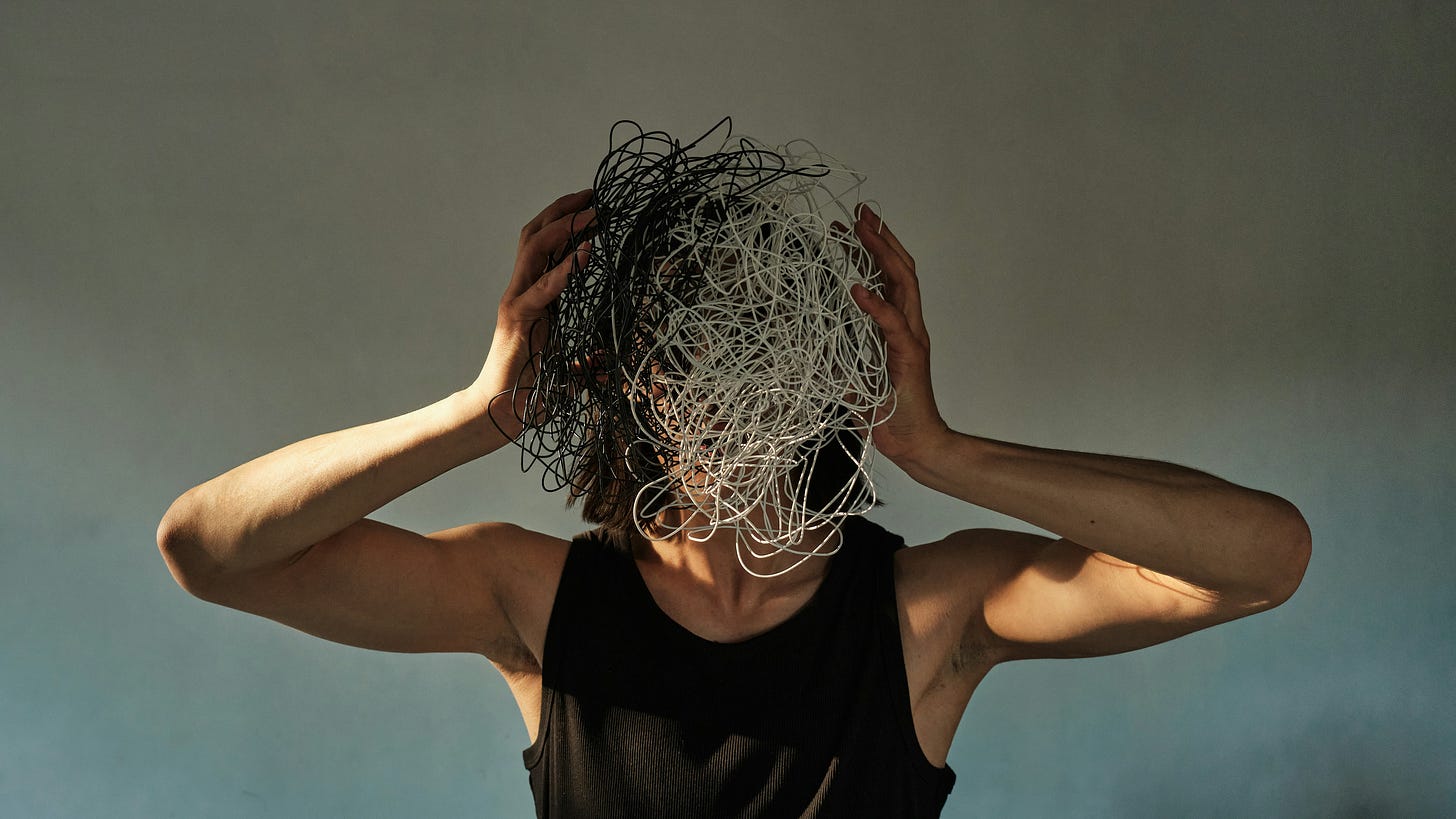 A person in a black tank top with their hands on their head. Their face is covered with a tangle of white and black wires. 