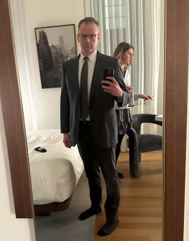 The author taking a mirror selfie in his (charcoal?) suit. The author's stunning wife is caught in passing in the background. 