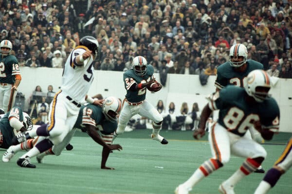 An action shot of two football teams. In the middle of the image a person is holding a football and running.