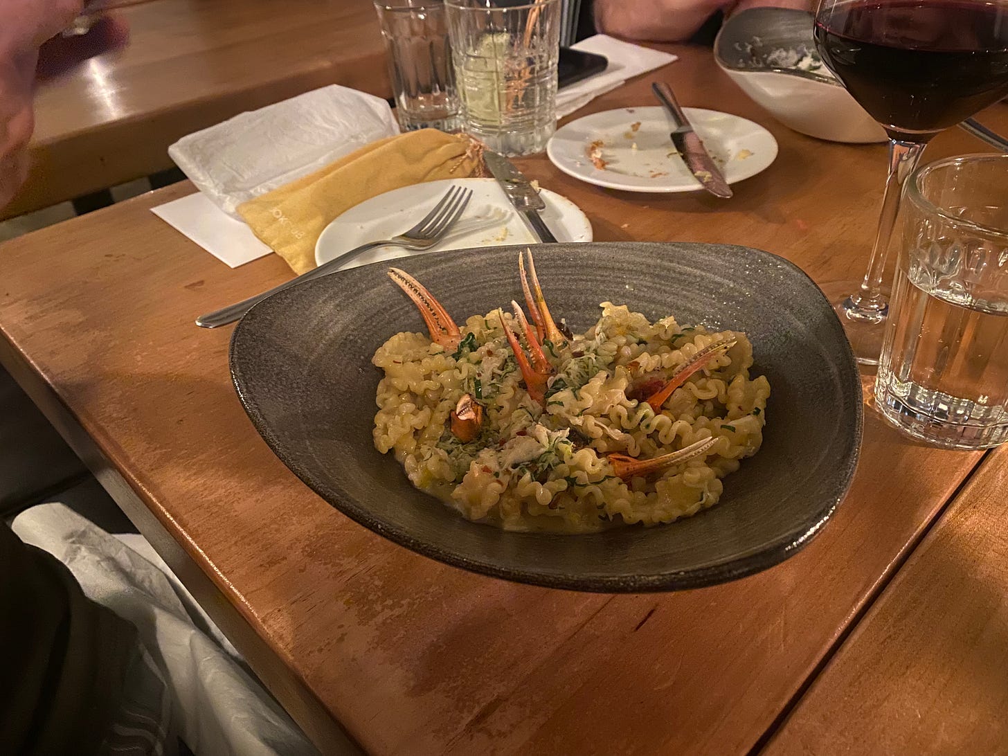 An asymmetrical bowl of mafalde noodles in a creamy sauce with greens. Small crab claws stick out from the pasta, five or six of them. Also on the table are glasses of water and side plates with forks and knives resting on them.