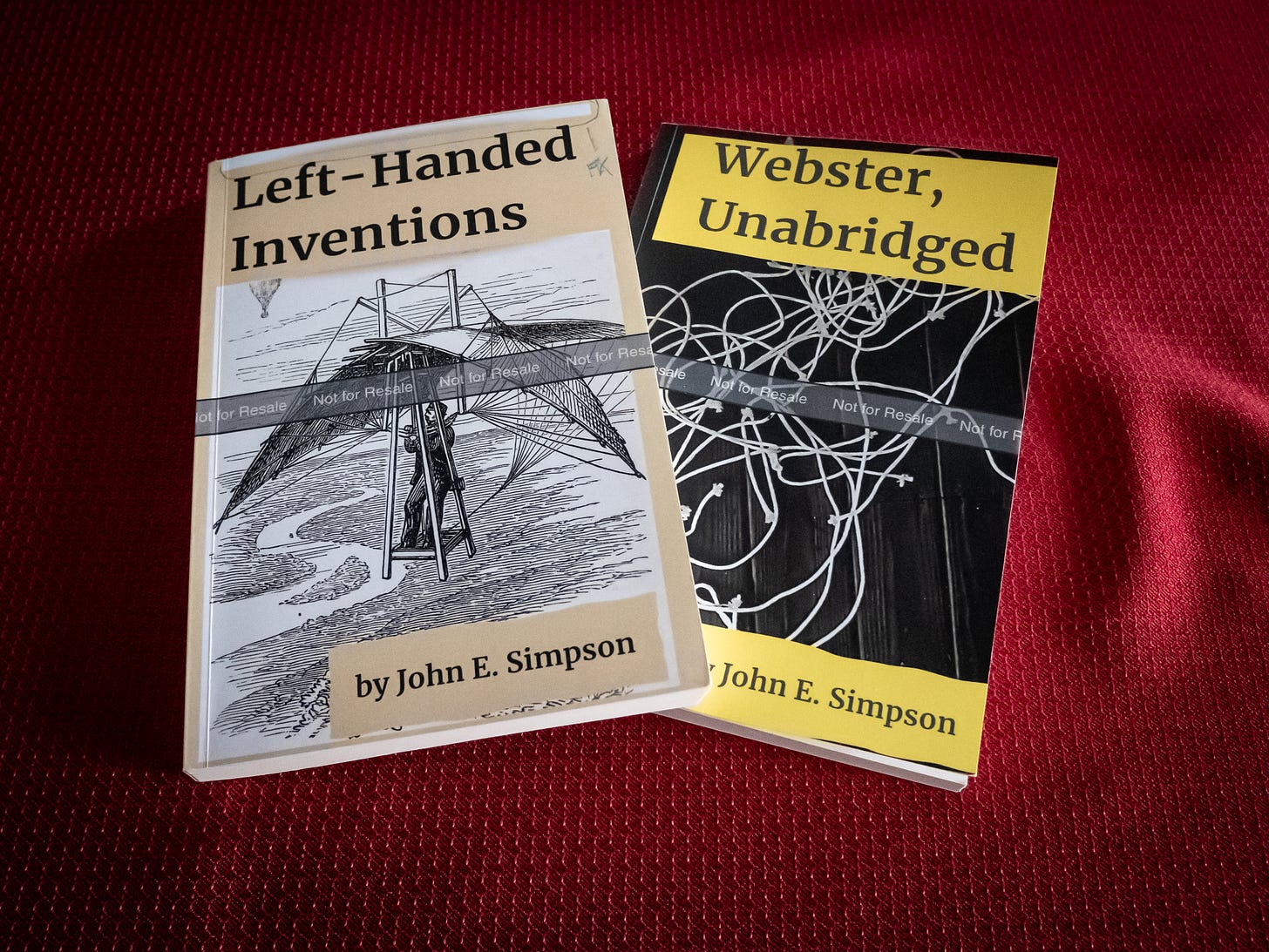 A photograph of two 6" x 9" paperback books by the author, lying on a table covered with red cloth. One book is called 'Left-Handed Inventions'; the other, 'Webster, Unabridged.' Both book covers include a grey band running across the middle, labeled "Not for Resale."