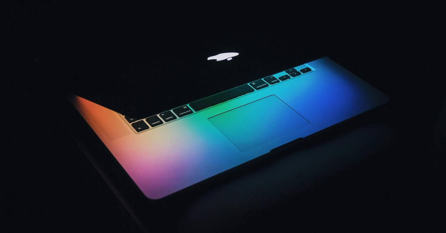 A laptop is mostly closed in a darkened space. Rainbow light from the screen, not visible, plays over the keyboard.