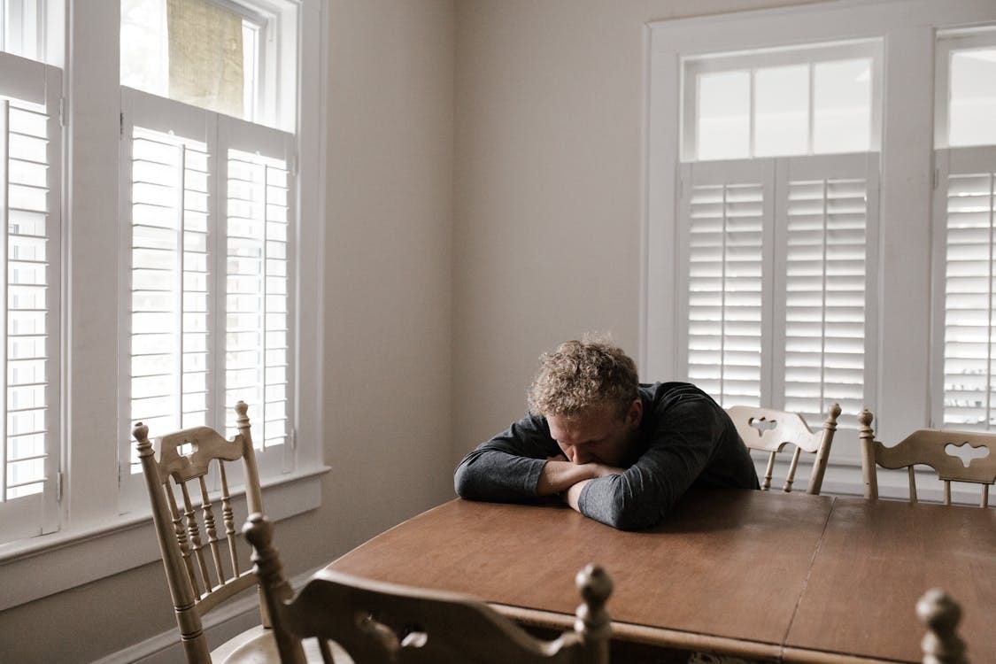 Free An adult man with emotions of sadness and anxiety sitting alone at a wooden table near windows. Stock Photo