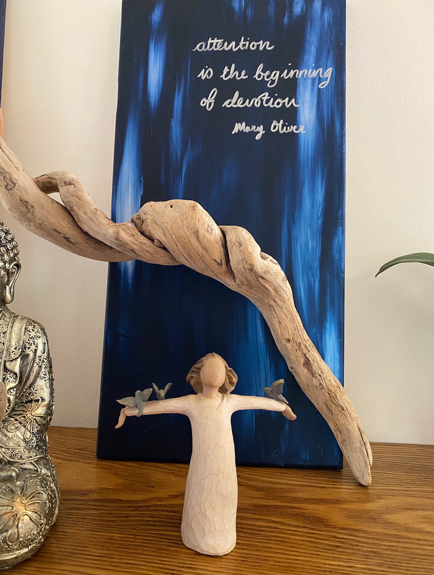 a statuette of a woman with arms outstretched holding blue birds on each arm, beneath a blue canvas with the words "attention is the beginning of devotion Mary Oliver" written on it. A sliver of a silver seated Buddha is visible to the left of the image