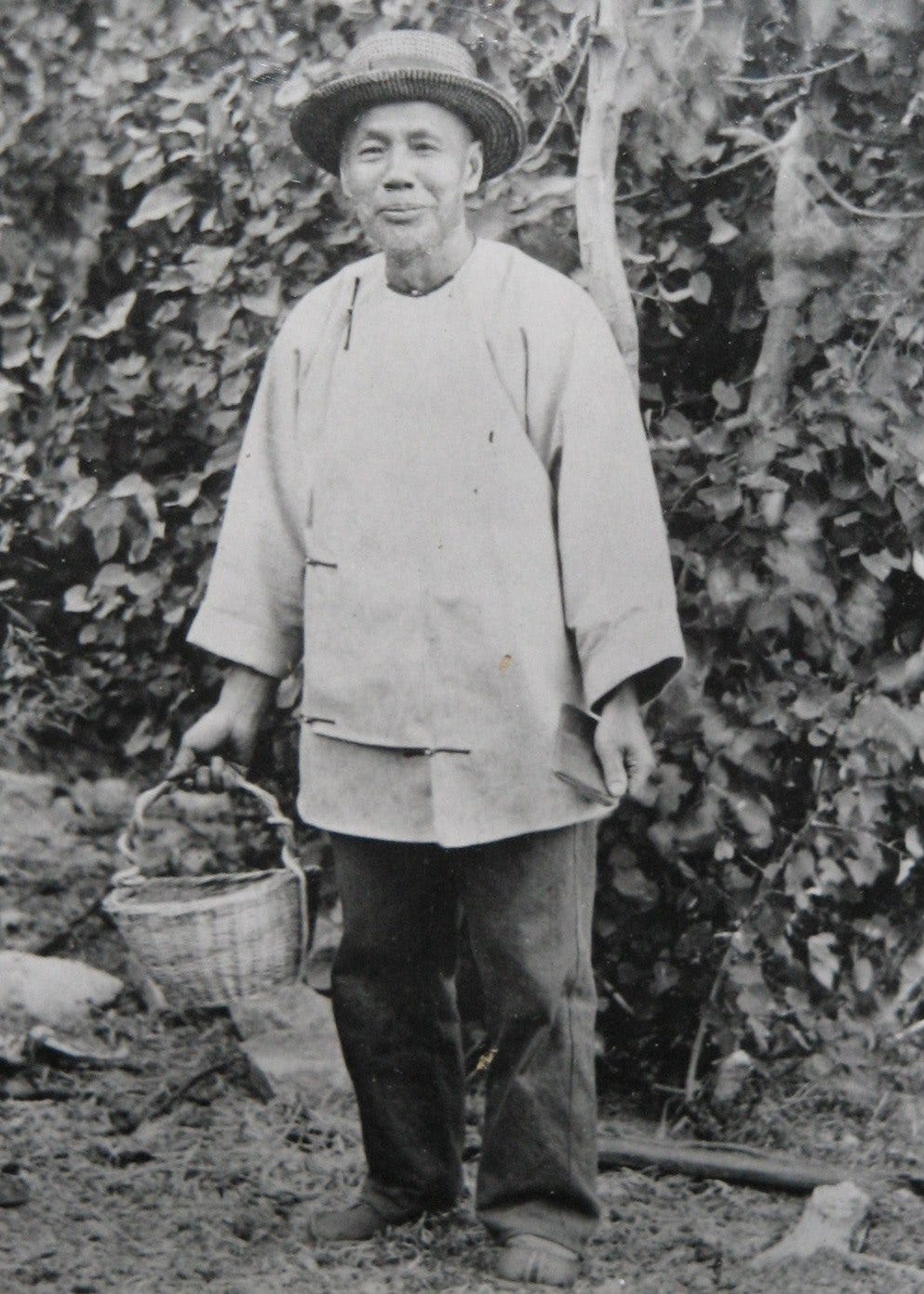 A black and white old photo with a middle-age looking Asian man standing in the field, holding a basket and smiling at camera.