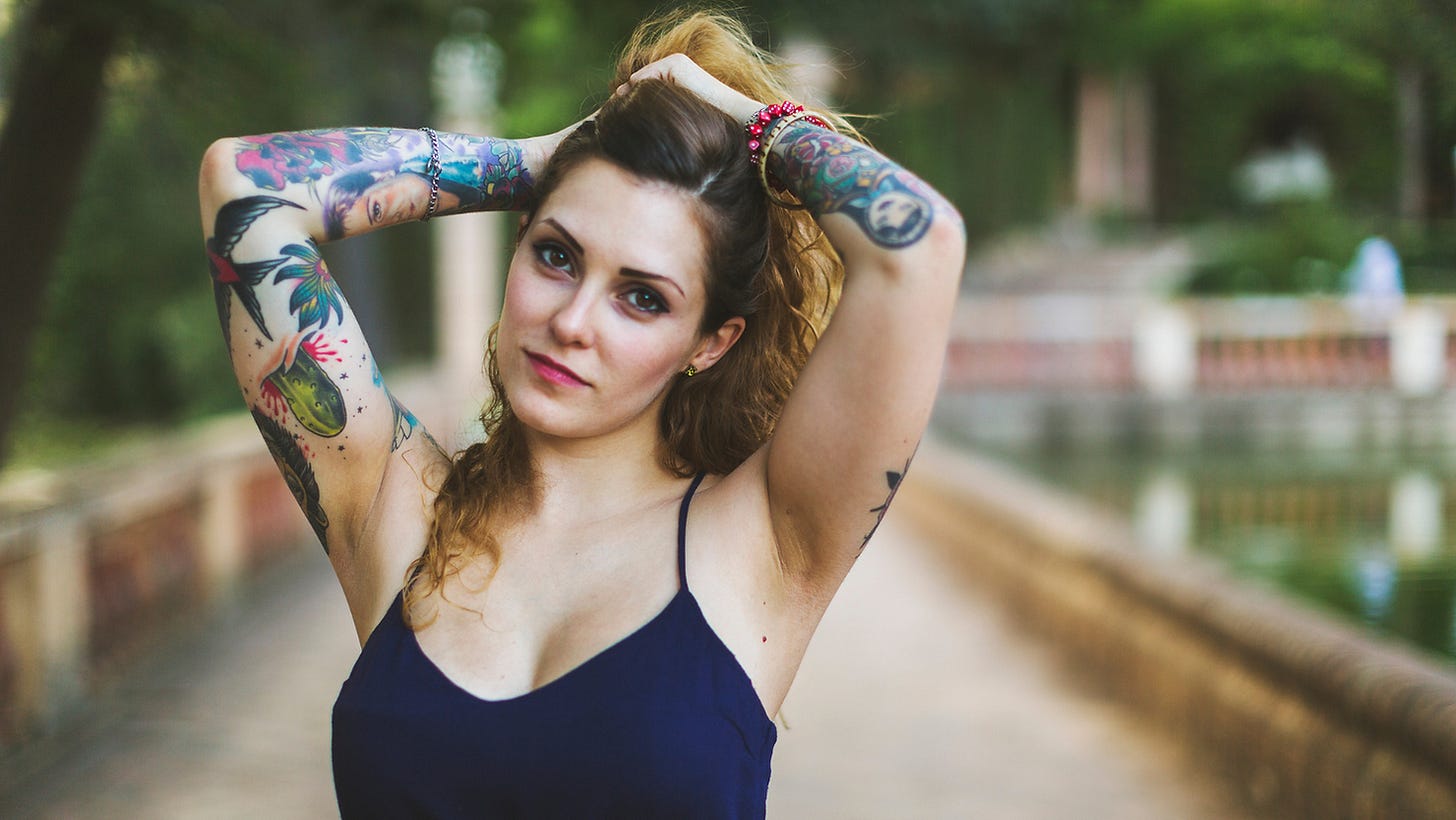 A tattooed white woman in a blue vest top holding her hiar