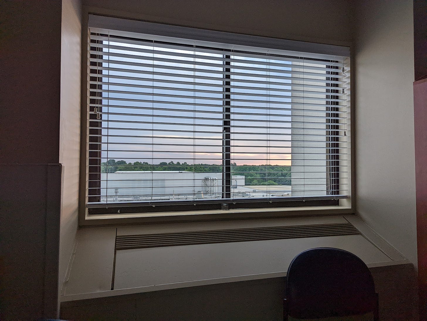 A window with blinds and a built-in AC unit. The view is of industrial HVAC equipment on a roof painted white, a tree line in the distance, and the sun rising through some clouds.