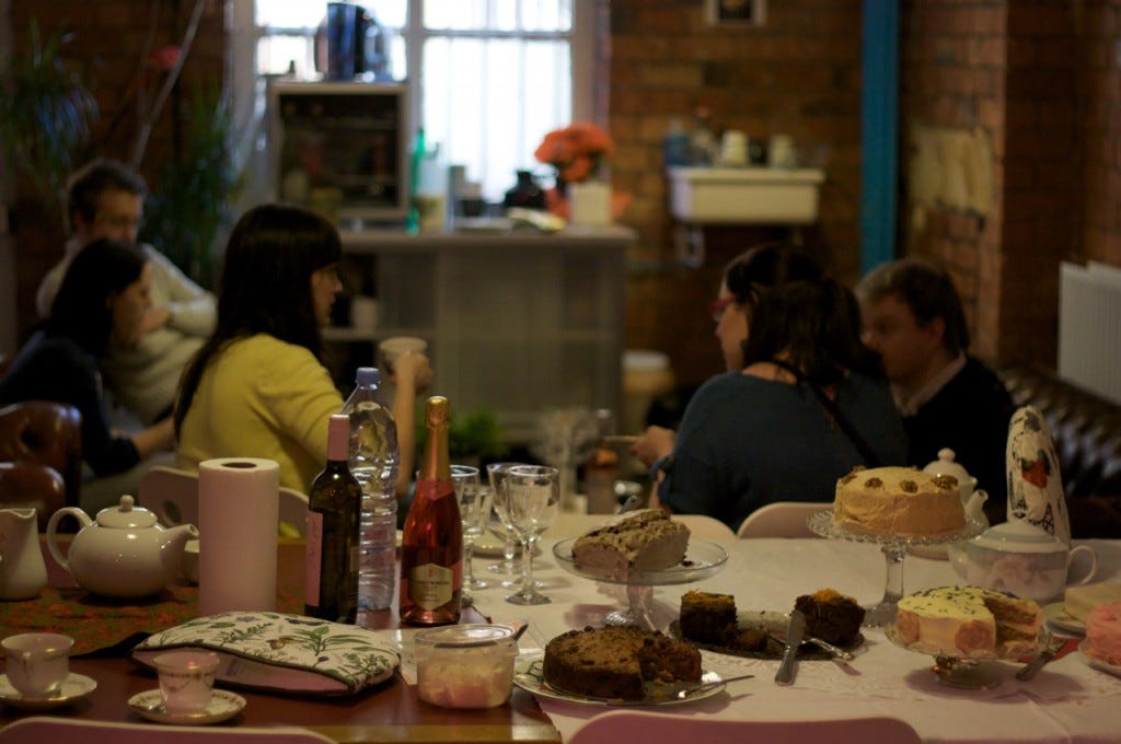 Group of people sat by a table eating cake and drinking tea.