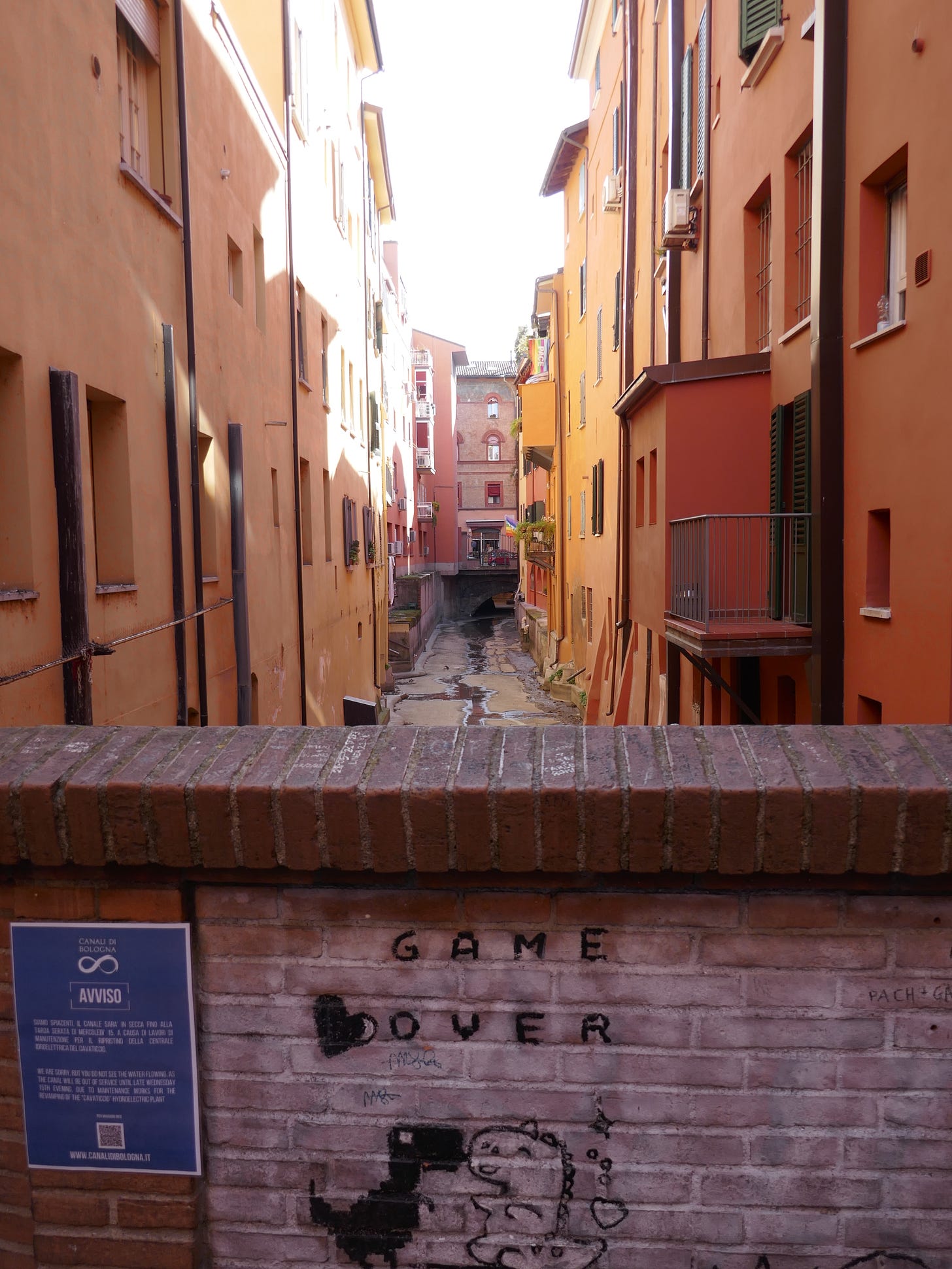 A view of Bologna's hidden canal, with a brick wall showing street art of two dinosaurs kissing with a heart and the words "Game Over."