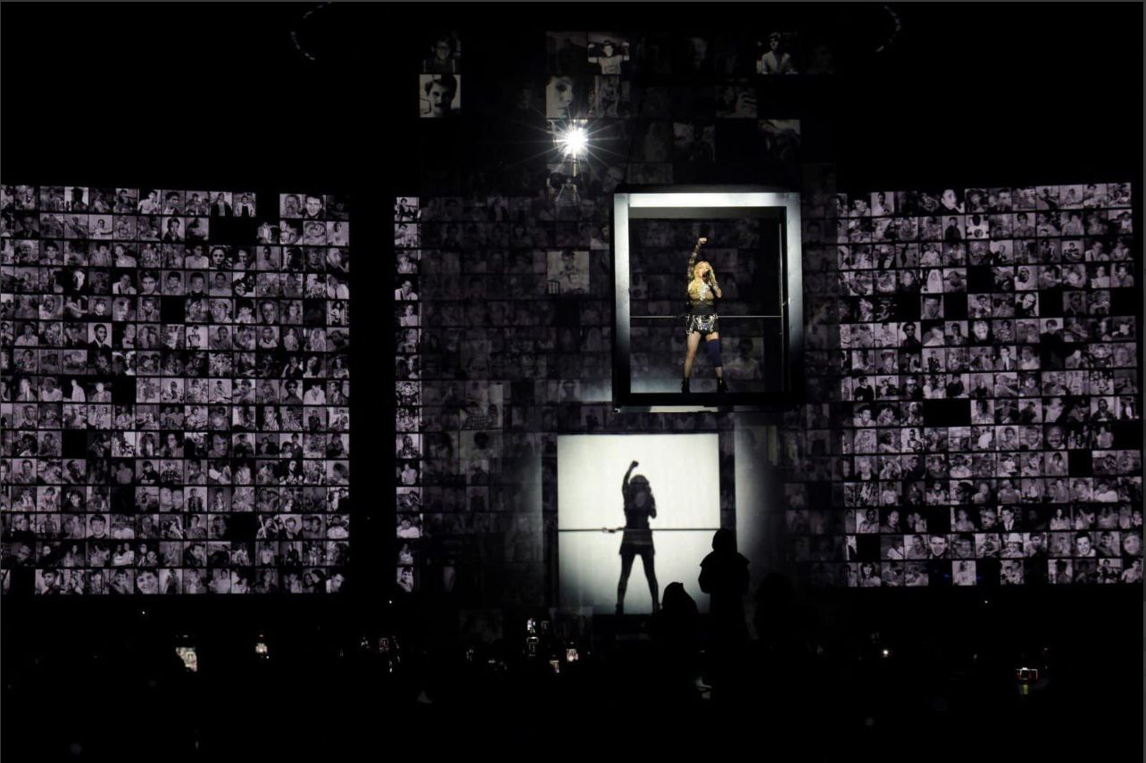 Madonna, suspensa em uma espécie de elevador, à frente de telões imensos com milhares de fotos de vítimas do HIV.