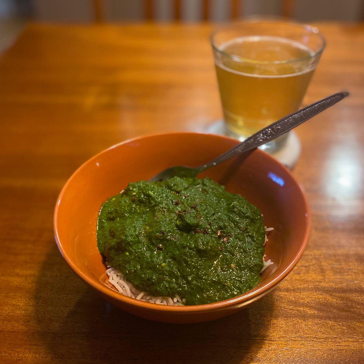 An orange bowl of basmati rice with dark green saag paneer overtop, dusted with chili flakes. A fork sits at the back of the bowl, and a glass of beer is on the coaster behind it.