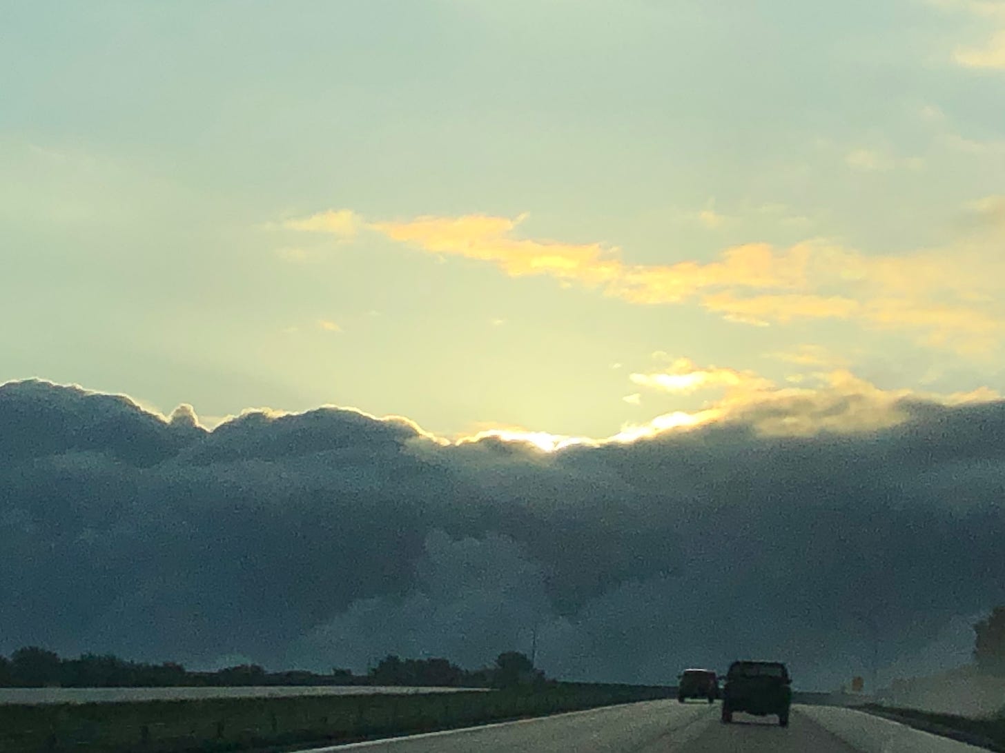 The sky is pale blue with brilliant gold clouds over a stretch of freeway. Between them, a continuous, opaque wall of dark gray clouds looms up, blocking out the rising sun.