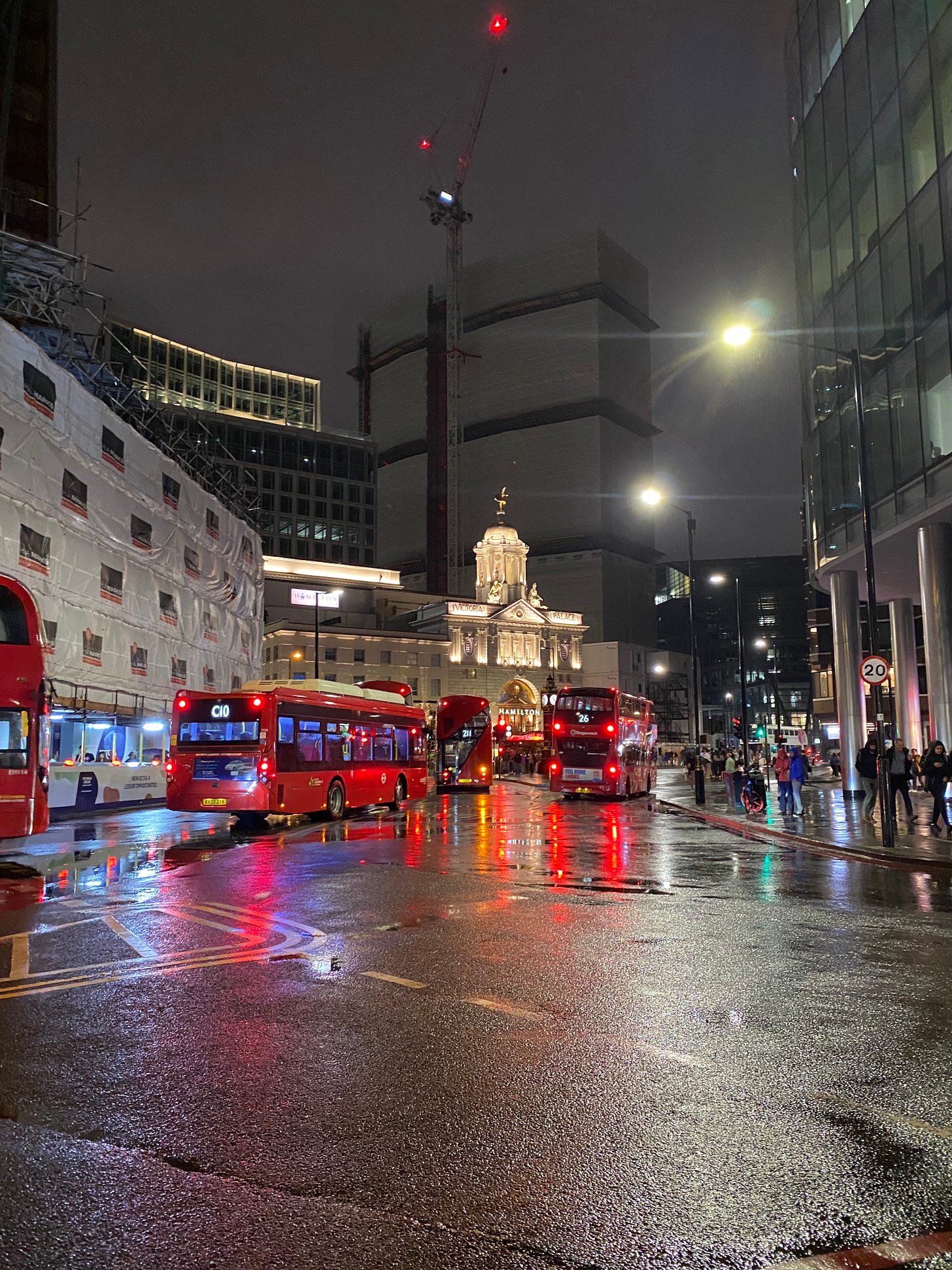 a theatre building lit up in white against a charcoal sky with red busses in a rainy street in the foreground